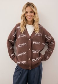 Bruine cardigan met herhaaldelijk "BIZUU" logo in het wit, met een V-hals, vijf knopen en geribbelde manchetten en zoom. Gecombineerd met losse blauwe broeken.