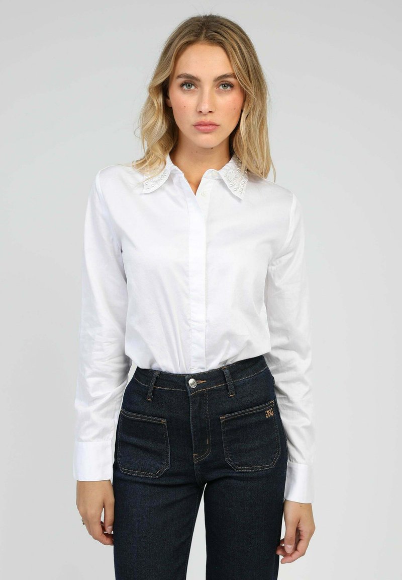 Chemise blanche à manches longues avec un col boutonné ornée de perles, associée à un jean taille haute bleu marine. Fond uni.