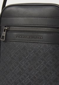 Black synthetic crossbody bag featuring a geometric pattern, zippered closure, and embossed logo on the front panel.
