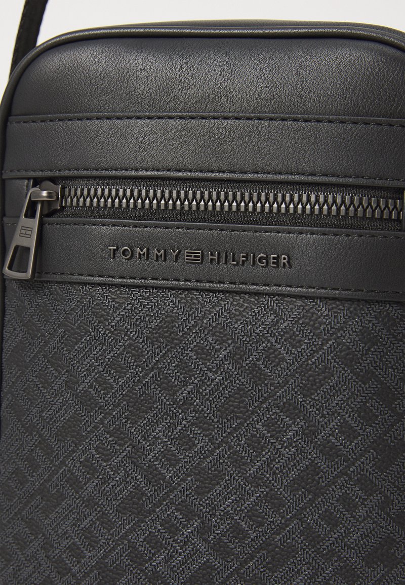 Bolso bandolera sintético negro con un patrón geométrico, cierre con cremallera y logo en relieve en el panel frontal.