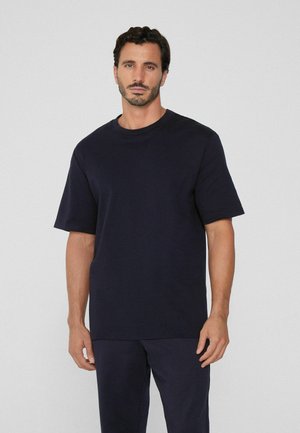 Mann mit kurzen dunklen Haaren und Bart, der ein einfaches schwarzes, übergroßes T-Shirt und schwarze Hosen vor einem schlichten grauen Hintergrund trägt.
