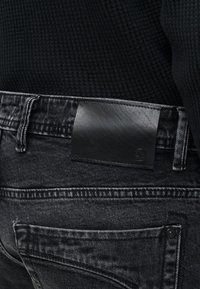 Κοντινό πλάνο στη μέση μαύρου τζιν με μαύρο δερμάτινο λογότυπο στο πίσω μέρος και μαύρο υφαντό πουλόβερ μπουκαρισμένο μέσα.