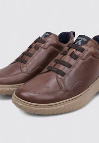 Zapatillas deportivas de cuero marrón con un acabado liso, que presentan acentos de cordones en marrón oscuro y una suela de goma con un patrón texturizado.