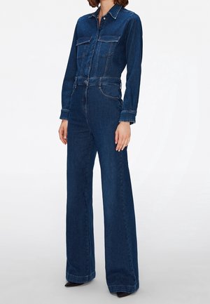 Person wearing dark blue denim jumpsuit with long sleeves, button front, chest pockets, and wide-leg pants with black pointed shoes.