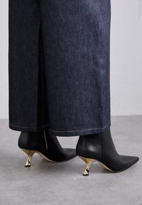 Bottes à chevilles en cuir noir avec des pointes pointues, dotées de talons aiguilles dorés. Portées avec un jean foncé, montrant une coupe nette à l'ourlet.