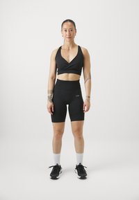 Crna sportska odjeća koja se sastoji od skraćenog crossover sportskog grudnjaka i visokih biciklističkih šortseva. Model nosi crne tenisice i bijele kratke čarape.