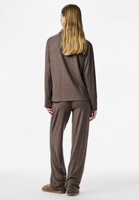 Žena s dugom plavom kosom, odjevena u labavu smeđu majicu dugih rukava i široke hlače u istoj boji, stoji okrenuta leđima.