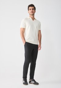 Cream polo shirt with a v-neck and short sleeves, paired with black slim-fit trousers and black sneakers with white stripes.