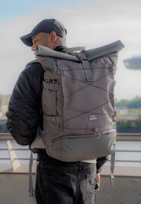 Mochila gris tipo roll-top con correas ajustables, tela texturizada y líneas de acento diagonales. Cuenta con un bolsillo frontal con cremallera y correas laterales ajustables.