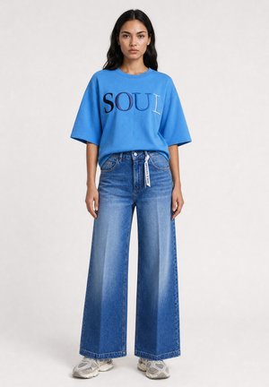 Woman stands wearing blue oversized t-shirt with "SOUL" text, wide-leg blue jeans, and white sneakers on light background.
