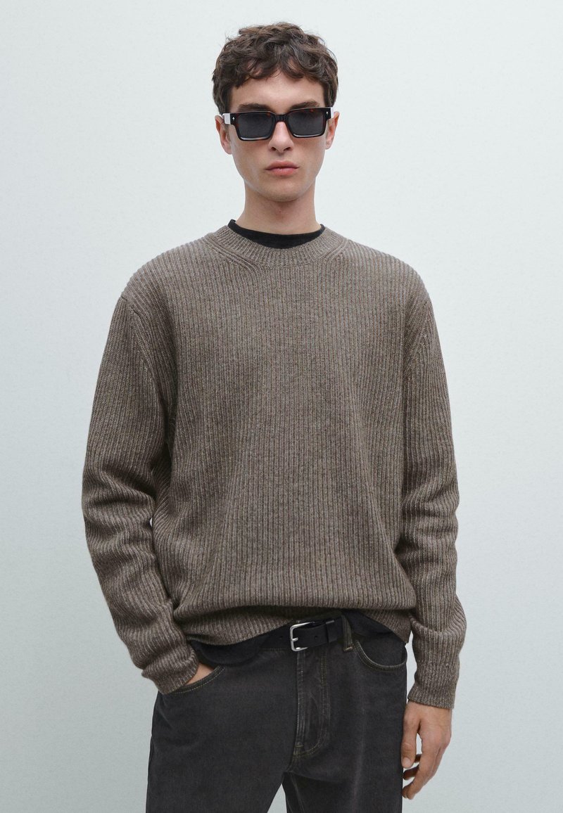Maglione a coste marrone con collo rotondo, indossato su una camicia nera, abbinato a jeans scuri e occhiali da sole neri rettangolari.