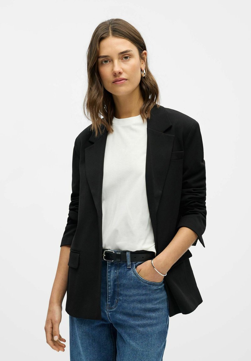 Žena s hnědými vlasy po ramena, která má stříbrné kruhové náušnice, černý blazer přes bílou košili, modré džíny a černý opasek, s rukou v kapse.