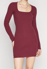 Burgunderfärgad ribbad stickad klänning med fyrkantig halsringning, långa ärmar och en figurnära silhuett. Slät textur med horisontellt ribbmönster.