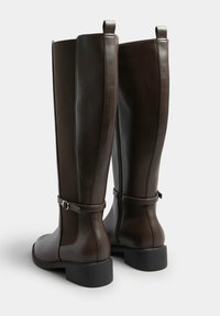 Brązowe skórzane botki do kolan z gładkim wykończeniem, panelami elastycznymi po bokach oraz regulowanym paskiem z klamrą na kostce. Niski, blokowy obcas.