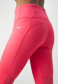 Ružové legíny s vysokým pásom vyrobené z elastického materiálu, so malým strieborným logom Nike a kontrastnými švami pre zvýraznenie detailov.