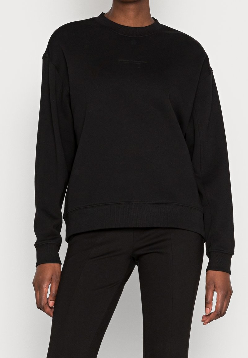 Personne portant un sweat-shirt noir uni à manches longues et un pantalon noir, debout devant un fond gris clair.