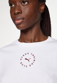 Valge puuvillane T-särk, millel on ümmargune burgundia punane logo, kus on kirjas "HYROX TRAINING CLUB" ja keskel puma siluett.