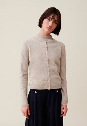 Cardigan beige chiaro con colletto alto, chiusura a sette bottoni e dettagli a costina ai polsini e all'orlo. Indossato sopra una gonna plissettata scura.