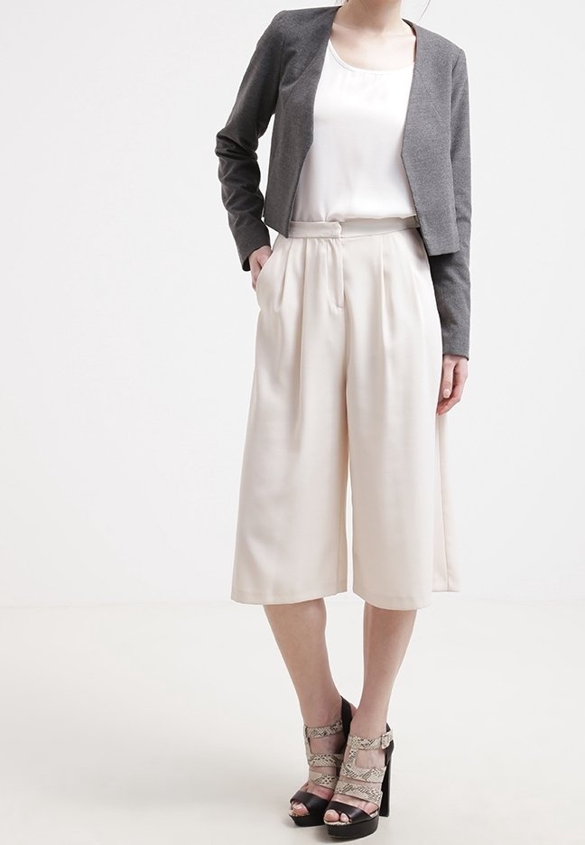 Femme portant un haut blanc sans manches, un blazer gris court, des culottes larges beiges et des sandales à talons avec des brides à imprimé serpent.