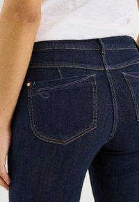 Jean en denim bleu foncé avec une coupe ajustée, présentant des coutures visibles, deux poches arrière et une texture lisse.