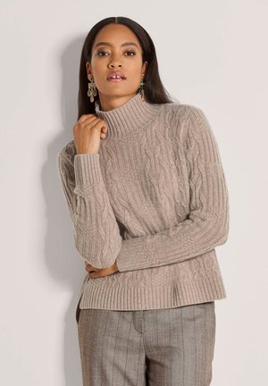 Vrouw met lang donker haar, gekleed in een beige kabelgebreide coltrui en gestreepte grijze broek, staand tegen een eenvoudige lichte muur.