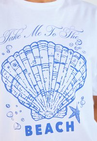 Vit t-shirt med en blå grafisk design av en sjöborre, bubblor och texten "Ta mig till STRANDEN" i fet blåa bokstäver.