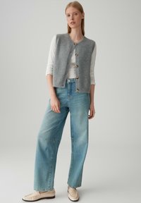 Gilet di lana grigia con apertura a bottoni, indossato sopra una camicia bianca a maniche lunghe. I jeans a gamba larga blu chiaro e le loafer beige completano l'outfit.