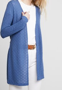Cárdigan azul de punto con un patrón texturizado, diseño de frente abierto y mangas largas. Combinado con una camisa blanca y un cinturón trenzado marrón.