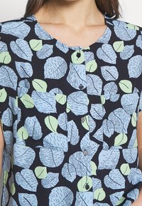 Camicia a maniche corte con bottoni, caratterizzata da uno sfondo blu navy e un motivo di foglie multicolore in blu chiaro e verde. Texture del tessuto morbida.