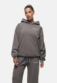 Felpa grigia oversize con tasca frontale e testo ricamato; realizzata in tessuto morbido e texture; abbinata a pantaloni della tuta coordinati.