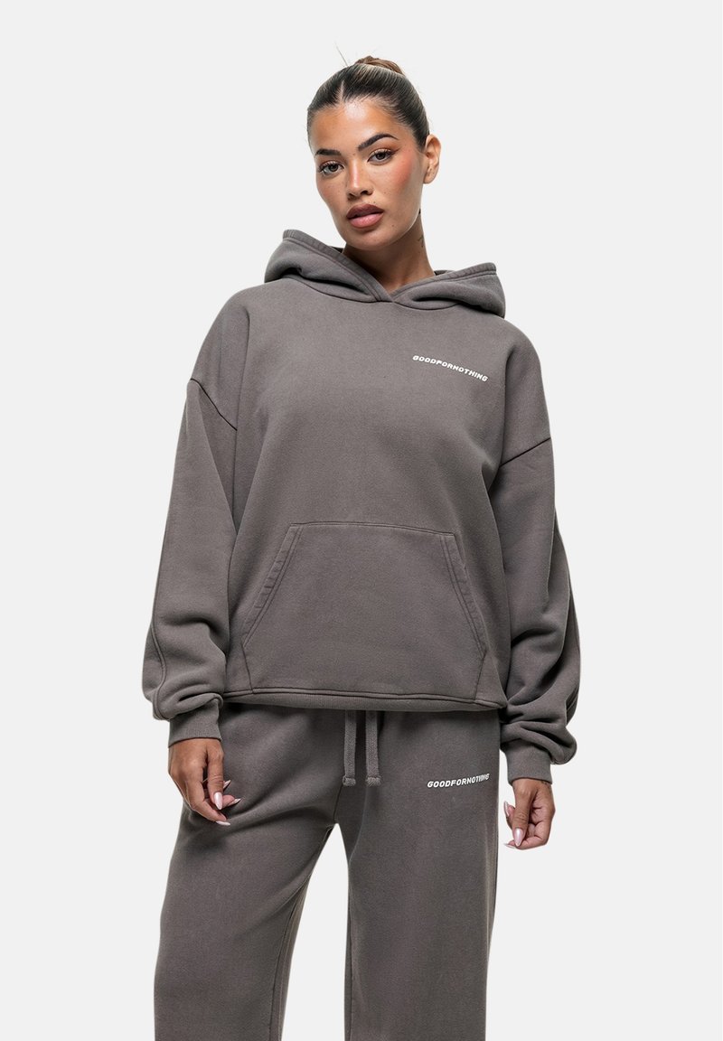 Felpa grigia oversize con tasca frontale e testo ricamato; realizzata in tessuto morbido e texture; abbinata a pantaloni della tuta coordinati.