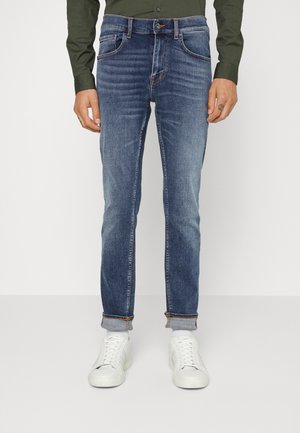 Homme portant un jean en denim bleu retroussé, des baskets blanches et une chemise boutonnée vert foncé, debout devant un fond uni.
