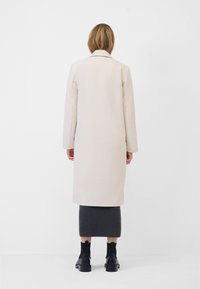 Lange crèmekleurige jas met een gestructureerd visgraatpatroon, enkelvoudig geknoopt ontwerp en een puntige kraag, gecombineerd met een grijze rok en zwarte enkellaarzen.