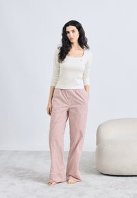 Bijela bluza s dugim rukavima u kombinaciji s crveno-bijelim širokim hlačama na pruge. Model stoji bos na svijetlosivoj pozadini.