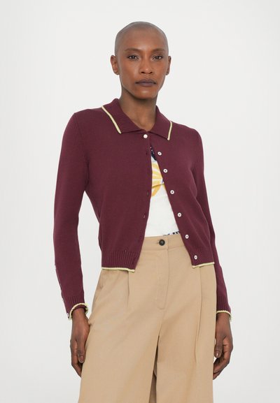 Cardigan bordeaux avec une texture côtelée, col et poignets jaunes contrastants, et fermeture à boutons. Porté par-dessus un haut imprimé et un pantalon crème.