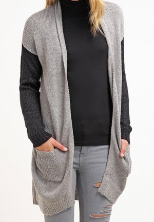 Grau gestrickte Strickjacke mit schwarzen Ärmeln, offener Vorderseite, zwei vorderen Taschen und lässiger Passform. Getragen über einem schwarzen Rollkragenpullover und ausgebleichten Jeans.