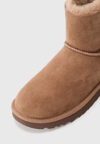 Bottine en suede marron avec un bout arrondi, dotée d'une doublure en polaire douce et d'une semelle en caoutchouc flexible et texturée. Détails de couture décorative présents.