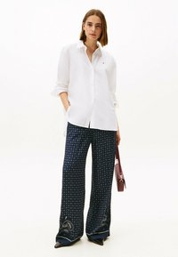 Camisa blanca de botones con las mangas arremangadas, combinada con pantalones anchos azul marino que presentan patrones redondos y de paisley, acentuada por un bolso oscuro.