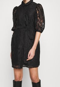 Abito in pizzo nero con silhouette aderente, chiusura frontale con bottoni, colletto e maniche a sbuffo. La vita legata accentua la forma.