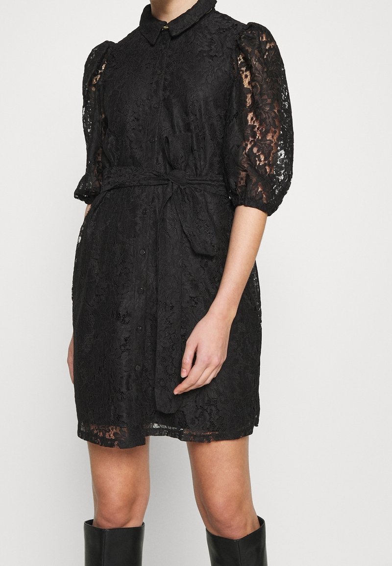 Abito in pizzo nero con silhouette aderente, chiusura frontale con bottoni, colletto e maniche a sbuffo. La vita legata accentua la forma.