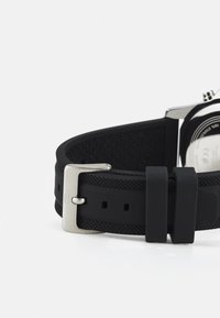 Schwarzes Gummiuhrband mit strukturiertem Design und silberner Metallschnalle. Verstellbare Länge mit mehreren Löchern zur Anpassung.