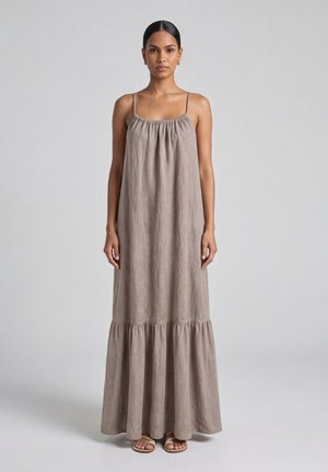 Donna con capelli scuri raccolti in uno chignon basso, indossa un abito lungo ampio taupe senza maniche con sottili spalline e sandali open-toe su uno sfondo grigio uniforme.