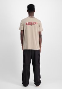 Beige bavlněné tričko s kulatým výstřihem, na zadní straně s červeným a bílým grafickým textem "ALPHA". Nosené s černými cargo kalhotami.