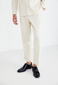 Pantalones de traje beige ajustados combinados con zapatos de cuero negros con cordones. El tejido parece suave, con líneas nítidas y un corte estructurado.