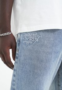 Jeans in denim azzurro chiaro con ricamo bianco della scritta "DON'T HARDY designs" sulla gamba inferiore, abbinati a una maglietta bianca semplice.