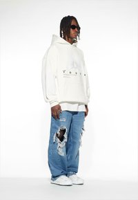 Felpa oversize bianca con stampa grafica, abbinata a jeans blu strappati con grandi strappi. Calzature: sneakers bianche. Occhiali da sole scuri indossati.
