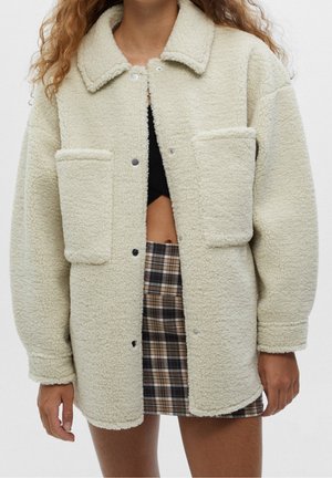 Femme portant une veste sherpa crème oversize avec de grandes poches avant, un crop top noir et une jupe à carreaux, debout devant un fond uni.