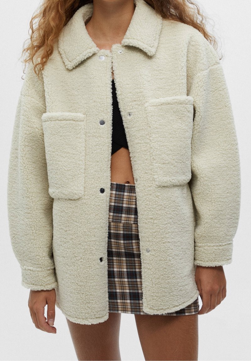 Femme portant une veste sherpa crème oversize avec de grandes poches avant, un crop top noir et une jupe à carreaux, debout devant un fond uni.