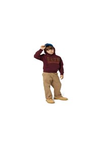 Felpa maroon con il logo "1973" e "Timberland", pantaloni beige e stivali tan. Cappellino blu indossato all'indietro, in posizione eretta su uno sfondo bianco.
