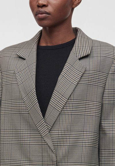 Personne portant un haut noir et un blazer à carreaux gris et noir avec de larges revers, visible des épaules jusqu'au milieu de la poitrine.